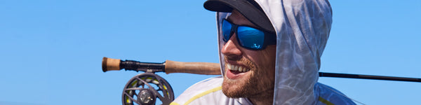 An angler wearing a light hoodie smiles as they balance their fly rod across their back.