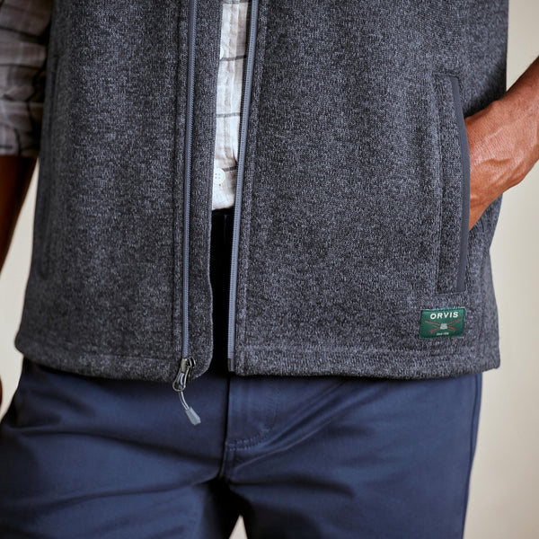 Person wearing a grey fleece vest with a brand logo on a neutral background