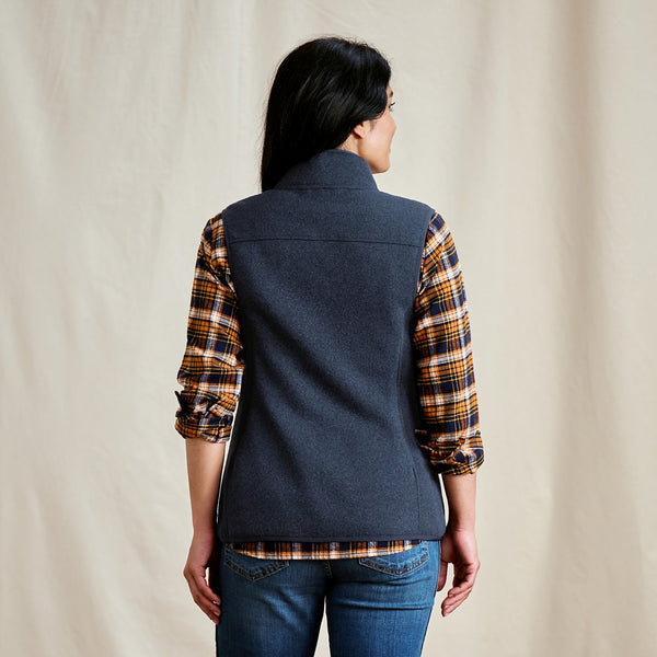 Person wearing a navy vest over a plaid shirt with a beige background