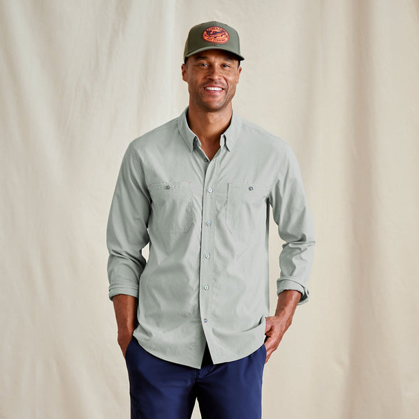 Man wearing a light grey button-up shirt and navy pants with a cap on a beige background