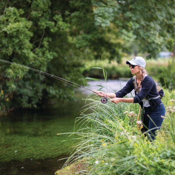 A person fly-fishing by the river, casting the fishing line into the water.