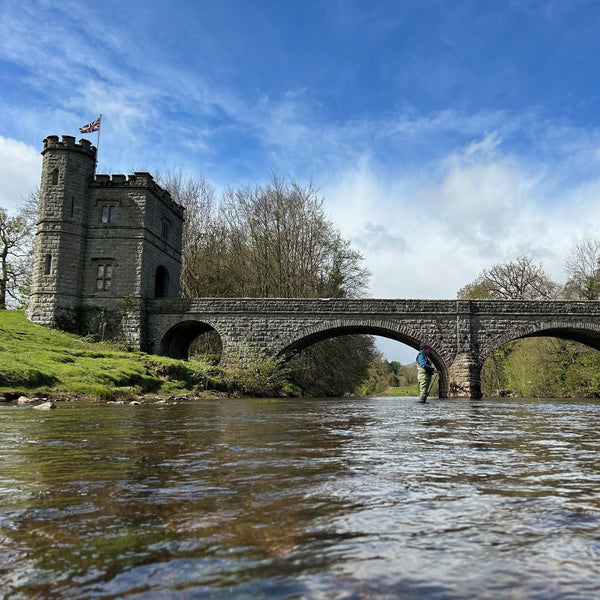 man fly fishing on river bed in front of bridge and castle