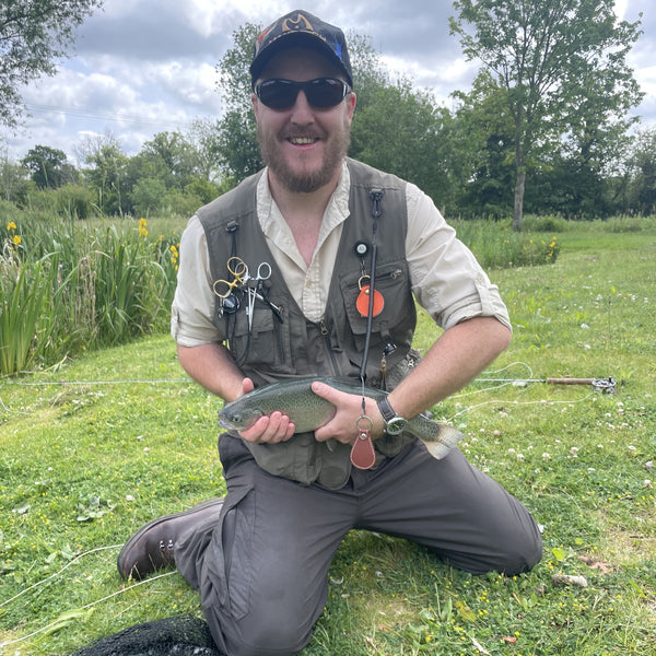 Man in fishing gear holding a fish outdoors on grass with trees in the background