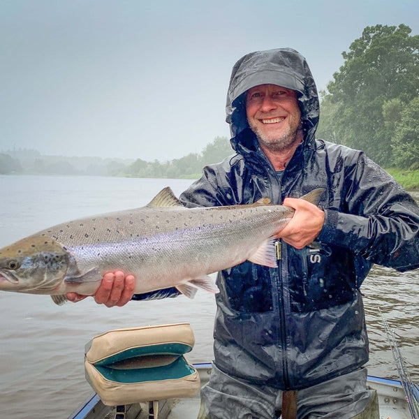 Man holding a large fish on a boat with a lake and trees in the background