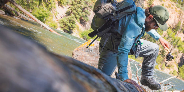 A fully-outfitted angler wearing waders hikes over a downed log in the river