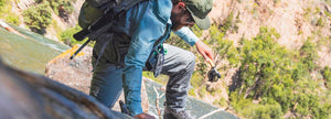An angler wearing waders climbs out of the water, over a fallen log