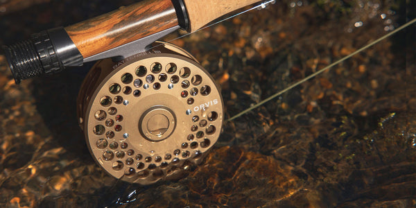 A close-up of a Battenkill Reel on an Orvis Superfine glass rod, sitting on the riverbed in shallow water