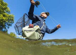 An angler wearing a waterproof hip pack casts their line whilst standing waist-deep in the river.