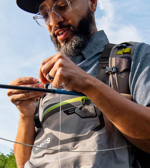An angler ties a knot while wearing waders and a ball cap
