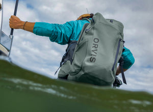 Person holding a large grey backpack with 'Orvis' branding above water.