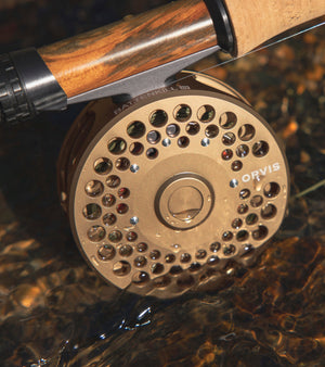 Close-up of a fly fishing reel with wooden handle on a reflective water surface.