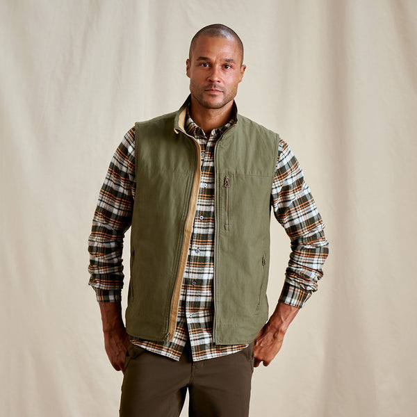 man wearing brown pants, shirt and green vest in front of beige background