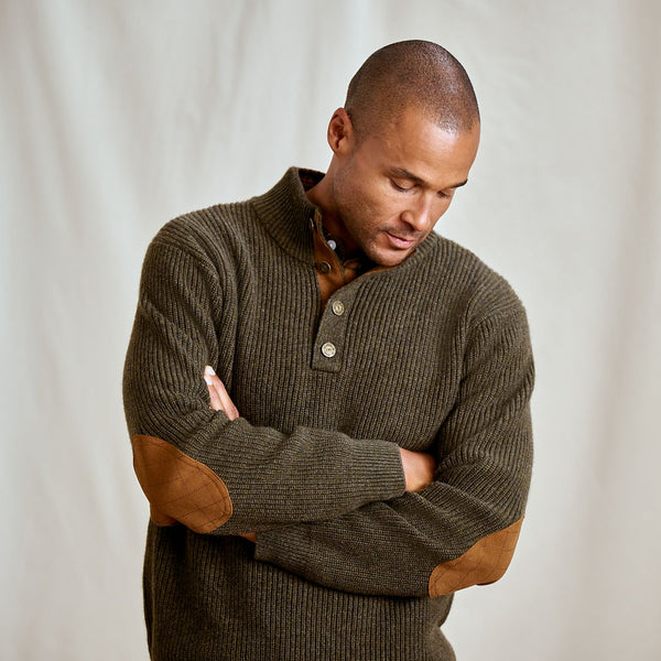Man wearing a brown ribbed sweater with leather elbow patches against a plain background