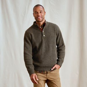 Man wearing a brown ribbed sweater with a plaid shirt underneath against a plain background
