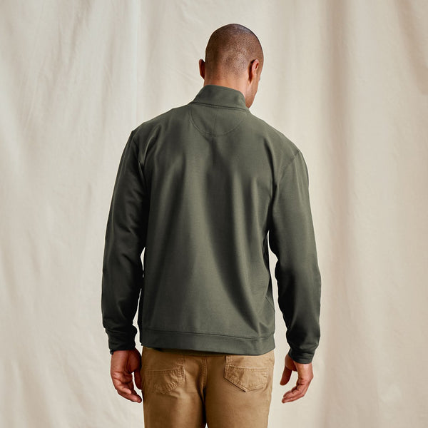 Man wearing a green half-zip sweatshirt and brown pants against a beige background