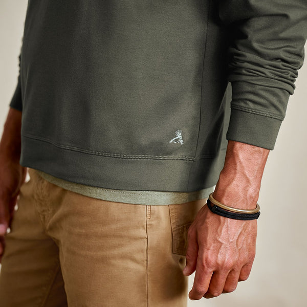 Person wearing a green sweatshirt with a small logo and beige pants, holding their arm.
