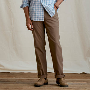 Person wearing brown pants and a plaid shirt on a wooden floor with a beige wall background