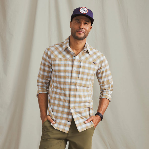Man smiling to the camera wearing blue fishing cap, beige and white checkered shirt and military green pants in front of beige background