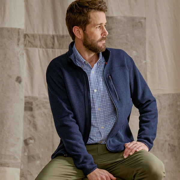 Man wearing a navy jacket over a checkered shirt sitting against a stone wall.