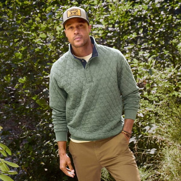 Man facing camera wearing fishing cap, dark green sweater and brown pants