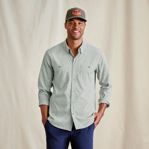 Man wearing a light grey button-up shirt and navy pants with a cap on a beige background
