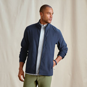 Man wearing a navy jacket over a light grey shirt with green pants against a beige background