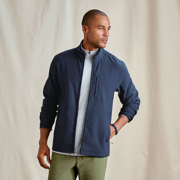 Man wearing a navy jacket over a light grey shirt with green pants against a beige background