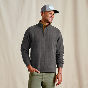 Man wearing a grey quilted pullover, green pants, and a cap against a beige background