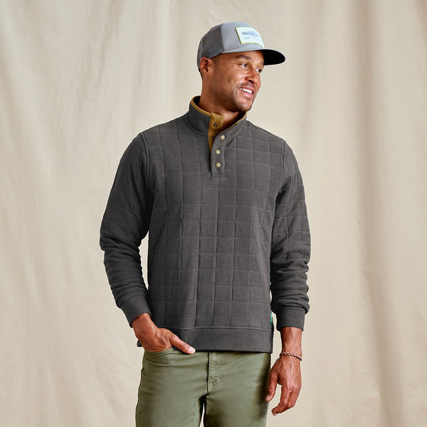 Man wearing a grey quilted pullover, green pants, and a cap against a beige background