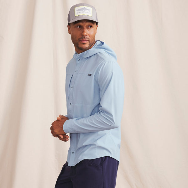 man in front of beige background wearing light blue jacket, blue pants and cap with fishing logo