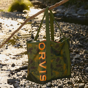 Green tote bag with 'Orvis' branding hanging on a branch by a stream.