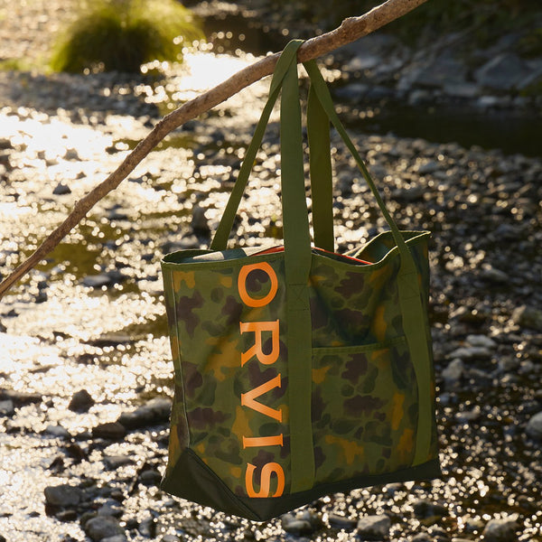 Green tote bag with 'Orvis' branding hanging on a branch by a stream.