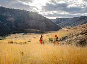 Man and woman stand in the mountains surrounded by hay on a wingshooting adventure