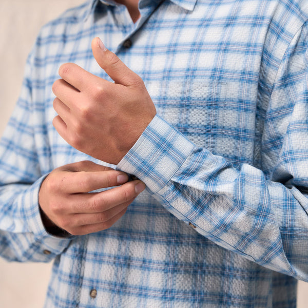 A close-up of a man wearing a light blue and white plaid long-sleeve button-down shirt, adjusting the cuff of the sleeve.