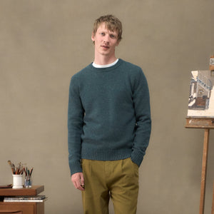 A man wearing a dark teal knit sweater and olive green trousers stands in a studio setting with an easel and painting desk visible in the background.