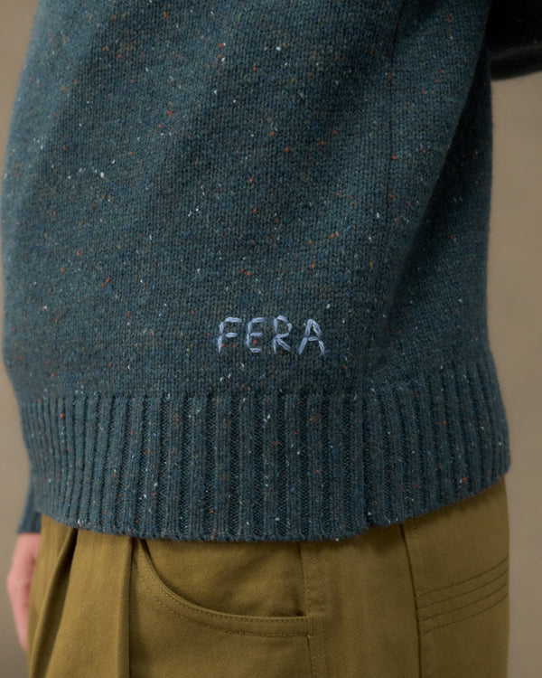 Close-up of a person wearing a dark teal speckled knit sweater with 'FERA' embroidered in white near the ribbed hem, paired with olive green trousers.