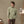 Load image into Gallery viewer, A man stands in a studio setting wearing a light sage green long-sleeve button-down shirt and brown trousers. The background features a warm beige wall, a wooden desk with art supplies, and an easel with a painting.

