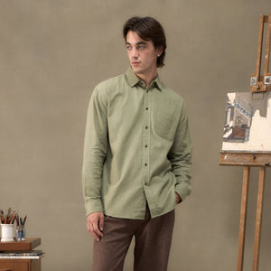 A man stands in a studio setting wearing a light sage green long-sleeve button-down shirt and brown trousers. The background features a warm beige wall, a wooden desk with art supplies, and an easel with a painting.
