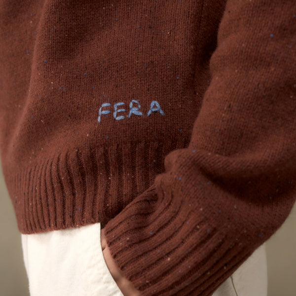 Close-up of a brown knit sweater with subtle multicolor flecks, featuring the word 'FERA' embroidered in light blue on the chest and ribbed cuffs, worn over white trousers.