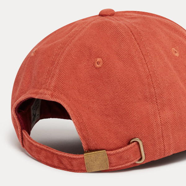 Close-up of the back of a textured terracotta orange baseball cap with an adjustable strap and brass buckle, on a light gray background.