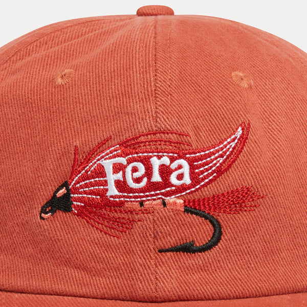 Close-up of a terracotta-colored baseball cap featuring an embroidered fly fishing lure with the word 'Fera' in white on the front.