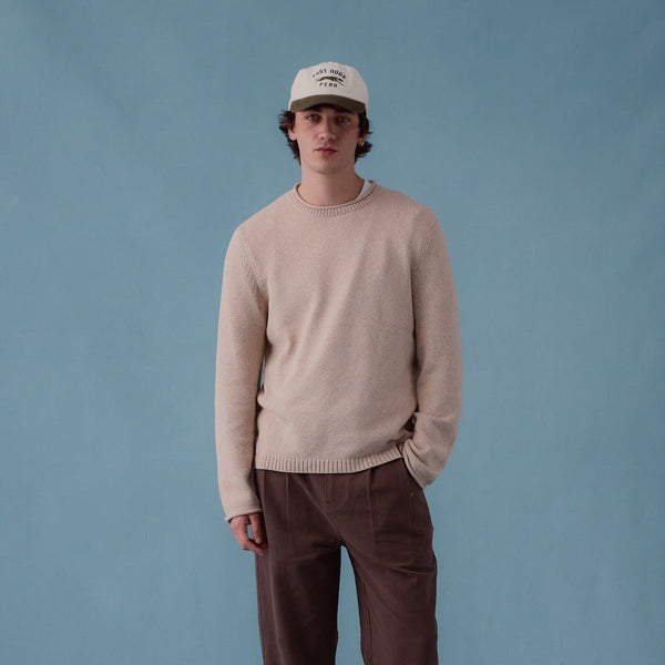 A man stands against a solid blue studio backdrop wearing a light beige crewneck sweater, dark brown corduroy pants, brown leather shoes, and a cream baseball cap.