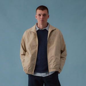 Man wearing a beige jacket, navy sweater, black shorts, and brown shoes against a blue background