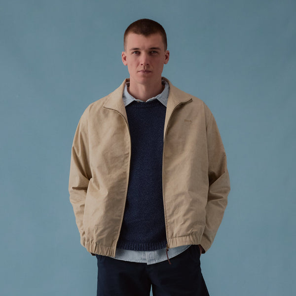 Man wearing a beige jacket, navy sweater, black shorts, and brown shoes against a blue background