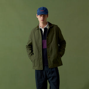 A man stands against an olive green studio backdrop wearing an olive green utility jacket over a dark navy sweater and white collared shirt, with dark blue jeans, brown boots, and a blue baseball cap.