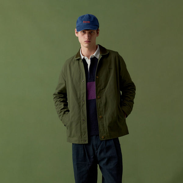 A man stands against an olive green studio backdrop wearing an olive green utility jacket over a dark navy sweater and white collared shirt, with dark blue jeans, brown boots, and a blue baseball cap.