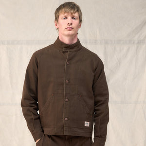 A man wearing a dark brown corduroy jacket with a high stand-up collar and matching brown pants, standing against a light beige fabric backdrop.