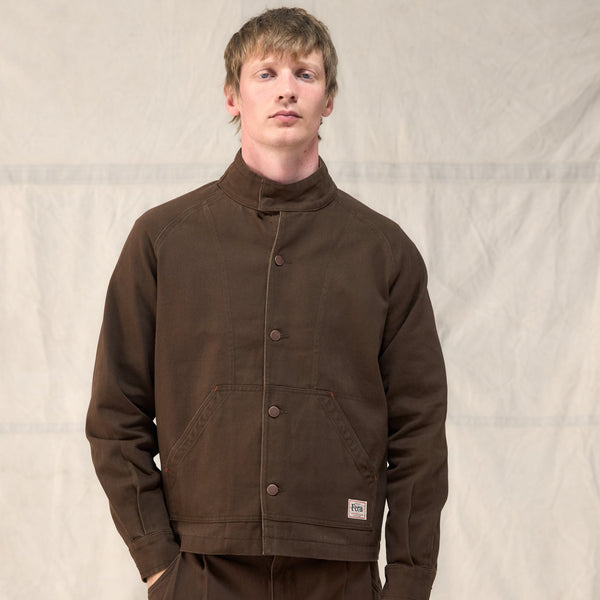 A man wearing a dark brown corduroy jacket with a high stand-up collar and matching brown pants, standing against a light beige fabric backdrop.