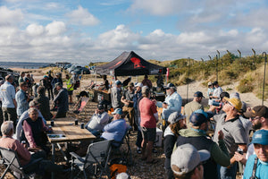 Group of people gathered around tables with a tent in a field under a cloudy sky