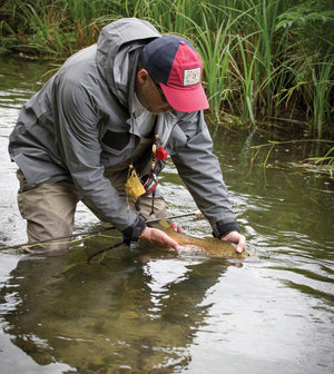 Man holds a fish in the water, wearing a red Orvis cap